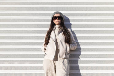 Fashionable teenage girl with hands in pockets standing while leaning on white wall - JRVF00298