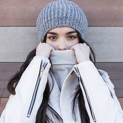 Beautiful teenage girl in knit hat covering her mouth with sweater against wall - JRVF00302