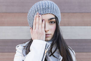 Teenage girl in knit hat covering her eyes with hands against wall - JRVF00304