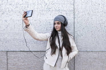 Teenager-Mädchen mit langen Haaren hören Musik durch Kopfhörer während der Einnahme selfie gegen silberne Wand - JRVF00309