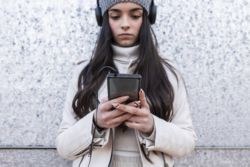 Teenager-Mädchen, das Musik über Kopfhörer hört, während es ein Telefon benutzt, das an einer silbernen Wand steht - JRVF00312