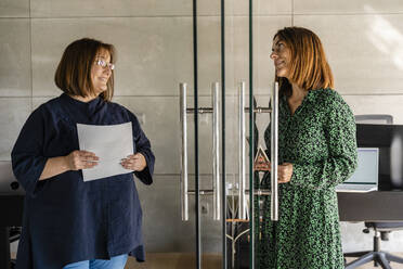Weibliche Mitarbeiter diskutieren, während sie an einer Glastür im Büro stehen - DLTSF01621