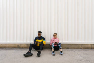 Smiling male and female athlete using mobile phone while sitting on floor against wall - EGAF01842