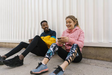 Male athlete looking at smiling woman using phone while sitting against wall - EGAF01843