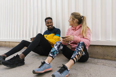 Smiling male and female athletes holding mobile phone while talking with each other against wall - EGAF01844