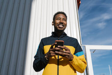 Smiling sportsman with mobile phone looking away while standing against wall - EGAF01853