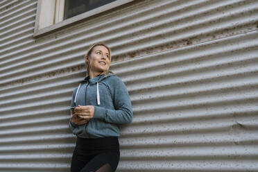 Lächelnde Sportlerin mit Mobiltelefon, die sich an eine Wand lehnt und wegschaut - EGAF01857