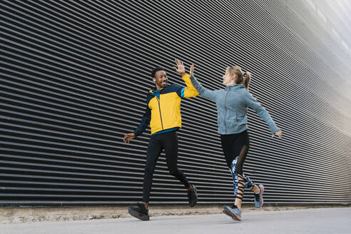 Männlicher und weiblicher Sportler machen High-Five beim Laufen auf dem Gehweg während des Sporttrainings an der Wand - EGAF01865