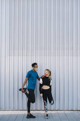 Male and female sportsperson with protective face mask doing warm up exercise while standing against wall during pandemic - EGAF01874