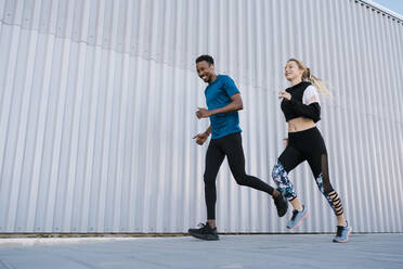 Smiling male and female athlete running on sidewalk by wall during sports training - EGAF01878