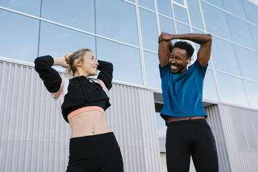 Female athlete looking at happy man while doing stretching exercise against glass building - EGAF01881