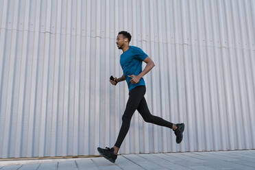 Male athlete with mobile phone running on sidewalk by wall during training - EGAF01890