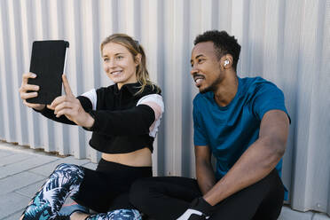 Junge Frau mit Mann nimmt Selfie durch digitale Tablet beim Sitzen auf dem Boden gegen die Wand - EGAF01896