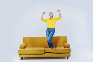 Fröhliche ältere Frau tanzt auf dem Sofa, während sie Musik über drahtlose Kopfhörer vor weißem Hintergrund hört - OIPF00370