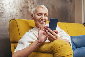 Cheerful senior woman using smart phone while sitting on sofa in living room - OIPF00377