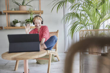 Smiling woman waving hand during video call while sitting at home - FMKF07040