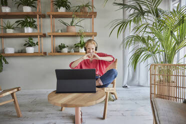 Happy woman making heart sign with hands during video call while sitting at home - FMKF07041