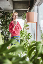 Nachdenkliche Frau mit Kaffeetasse inmitten von Pflanzen zu Hause - FMKF07046