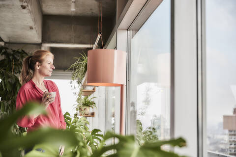 Frau mit Kaffeetasse schaut durch Glasfenster zu Hause, lizenzfreies Stockfoto