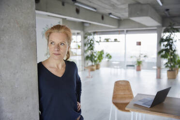 Serious woman with hand on hip leaning on wall at home - FMKF07052