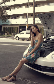Frau spricht in ihr Handy, während sie sich auf der Motorhaube eines Autos am Straßenrand abstützt - AJOF01124