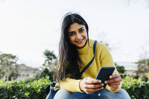 Lächelnde Frau mit Handy in der Hand, während sie in einem öffentlichen Park sitzt - XLGF01203