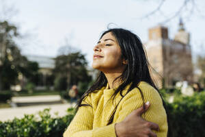 Smiling woman hugging self against sky in public park - XLGF01204