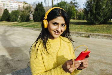 Lächelnde schöne Frau, die ein Mobiltelefon hält, während sie in einem öffentlichen Park über Kopfhörer Musik hört - XLGF01208