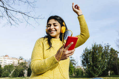 Lächelnde Frau mit Mobiltelefon tanzt beim Hören von Musik über Kopfhörer gegen den Himmel in einem öffentlichen Park - XLGF01210