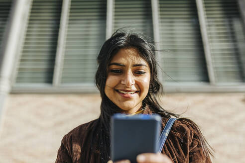 Lächelnde Frau, die ein Mobiltelefon gegen ein Gebäude benutzt - XLGF01219