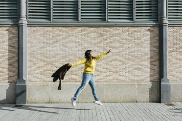 Woman with jacket jumping on sidewalk by wall - XLGF01222