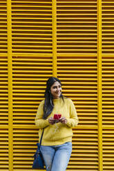 Lächelnde Frau mit Mobiltelefon, die über Kopfhörer Musik hört und wegschaut, während sie an einer gelben Wand steht - XLGF01224