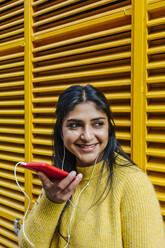 Lächelnde Frau, die ihr Smartphone in der Hand hält und wegschaut, während sie über Kopfhörer an der Wand Musik hört - XLGF01225