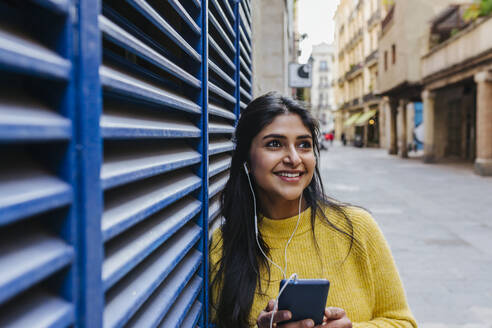 Lächelnde Frau mit Mobiltelefon, die wegschaut, während sie über Kopfhörer an einer blauen Wand Musik hört - XLGF01227