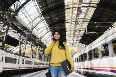 Lächelnde Frau mit Laptoptasche, die wegschaut, während sie auf dem Bahnsteig mit ihrem Smartphone telefoniert - XLGF01233