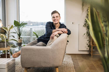 Smiling young man sitting on sofa against window in living room - UUF22749