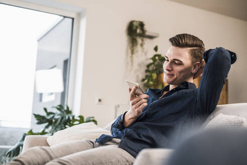 Entspannter junger Mann, der sein Mobiltelefon benutzt, während er zu Hause auf dem Sofa sitzt - UUF22754