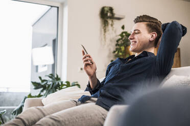 Fröhlicher junger Mann, der ein Mobiltelefon benutzt, während er zu Hause auf dem Sofa im Wohnzimmer sitzt - UUF22755