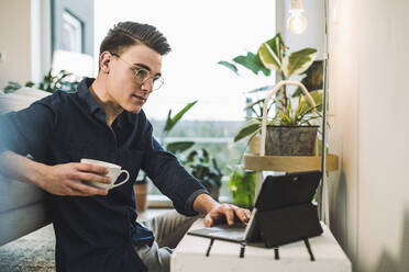 Junger Mann mit Brille, der einen Laptop benutzt, während er im Wohnzimmer sitzt - UUF22756
