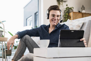 Smiling young man with wireless headphones watching video through digital tablet in living room - UUF22758