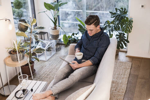 Junger Mann mit Kaffeetasse auf dem Sofa sitzend und mit digitalem Tablet im Wohnzimmer - UUF22761