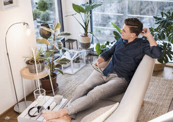 Young man with digital tablet looking away while sitting on sofa in living room - UUF22762