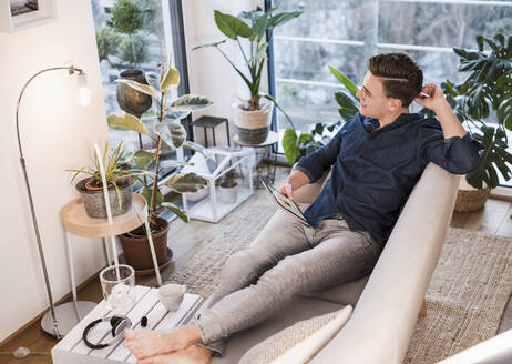 Junger Mann mit digitalem Tablet, der wegschaut, während er auf dem Sofa im Wohnzimmer sitzt - UUF22762