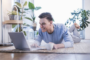 Lächelnder junger Mann, der einen Laptop benutzt und auf dem Teppich im Wohnzimmer liegt - UUF22763