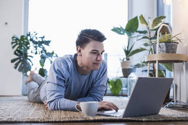 Junger Mann mit Kaffeetasse, der einen Laptop benutzt, während er auf dem Teppich im Wohnzimmer liegt - UUF22764