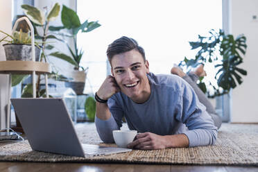 Glücklicher junger Mann mit Laptop auf dem Teppich im Wohnzimmer liegend - UUF22765