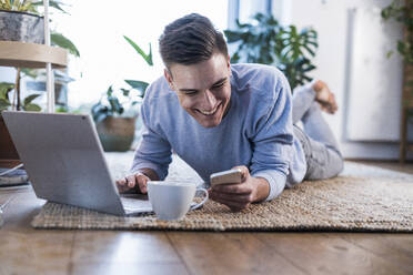 Fröhlicher junger Mann mit Laptop, der sein Smartphone benutzt, während er auf dem Teppich im Wohnzimmer liegt - UUF22768