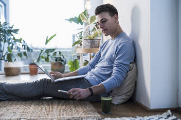 Young man with laptop checking smart phone while sitting in living room - UUF22771