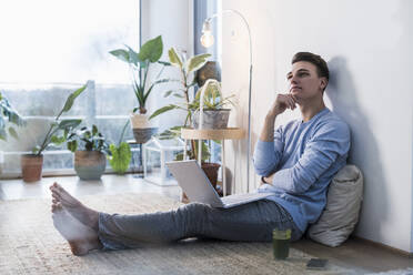 Nachdenklicher junger Mann mit Laptop, der im Wohnzimmer an der Wand sitzt - UUF22773