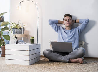 Junger Mann mit Laptop sitzt entspannt an der Wand im Wohnzimmer - UUF22775